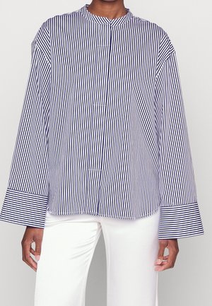 Femme portant un chemisier ample à rayures verticales bleu marine et blanches avec de larges poignets, associé à un pantalon blanc sur un fond uni.