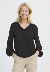 Smilende blond kvinde med hænderne i beige buksernes lommer, iført en løs sort bluse med lange ærmer, V-hals og knapdetalje.