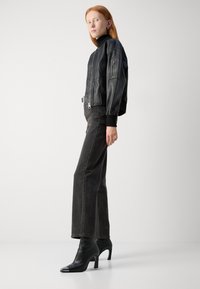 Chaqueta bomber de cuero negro con puños acanalados, cuello alto y bolsillos con cremallera, combinada con jeans rectos gris oscuro y botas de tacón negras.