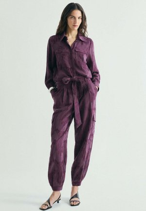 Mujer de pie con las manos en los bolsillos, con un mono morado estampado con cinturón y tacones negros de punta abierta sobre un fondo liso.