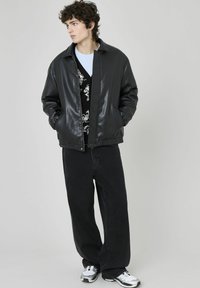Veste bomber en cuir synthétique noir avec une fermeture éclair, associée à un pull noir à motifs et un jean noir ample, complétée par des baskets grises.
