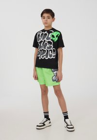 T-shirt nera con scritte bianche e verdi fluorescenti, abbinata a pantaloni corti verdi brillanti con un design grafico. Sneakers bianche e nere.