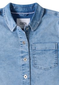Chemise en jean bleu clair avec boutons métalliques sur le devant, poche poitrine, et une étiquette blanche avec "CECIL" à l'intérieur du col.