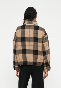 Beige und schwarzes Karo-Jackett mit Kragen, oversize Passform und strukturiertem Stoff. Besitzt weite Ärmel und einen geraden Saum.