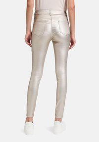 Jean skinny doré métallisé avec taille haute, comportant des poches arrière et une texture lisse et brillante, associés à des baskets blanches.