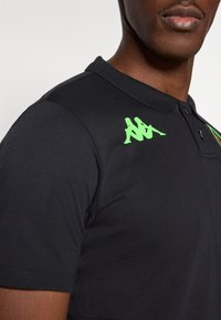 Fekete galléros póló rövid ujjakkal. A bal vállnál zöld logóval és sima, texturált anyagból készült.