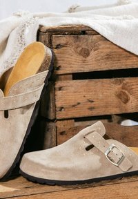 Lichtbeige suède klompen met gouden studs en een verstelbare zilveren gesp, leunend tegen een rustieke houten krat en een gestructureerde stof.