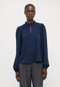 Blusa blu navy con maniche lunghe a sbuffo, colletto arricciato, apertura a occhiello e tessuto leggero e testurizzato. Abbinata a pantaloni grigi.
