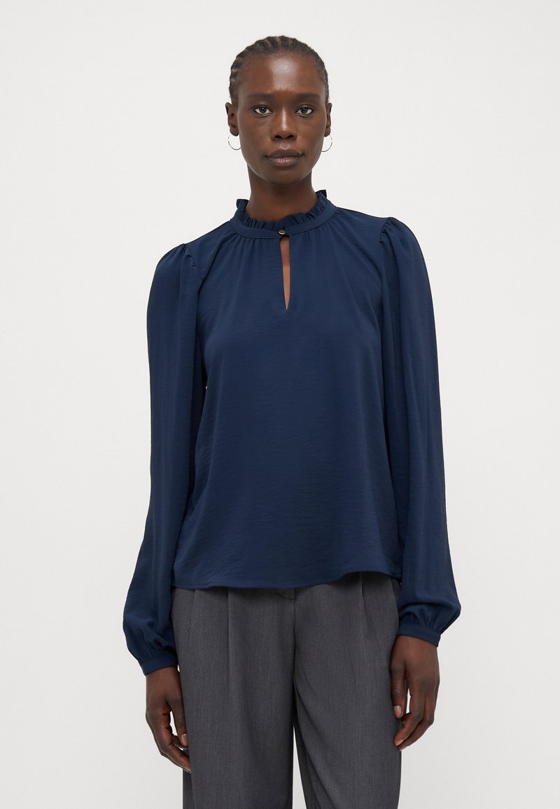 Blusa blu navy con maniche lunghe a sbuffo, colletto arricciato, apertura a occhiello e tessuto leggero e testurizzato. Abbinata a pantaloni grigi.