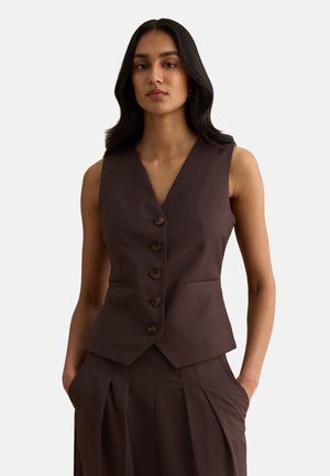 Frau trägt ärmellose dunkelbraune, geknöpfte Weste und passende hochtaillierte, plissierte Hose mit Händen in den Taschen, neutraler Hintergrund.