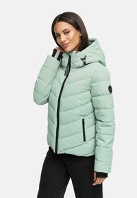 Woman wearing light green quilted hooded jacket with black zipper and pockets, posing with hand near neck, against white background.