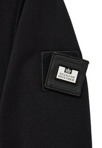 Un material de hanorac negru cu un patch pătrat negru pe care este inscripționat cu alb "WEEKEND OFFENDER" și un emblema cu o coroană. Suprafață simplă, texturată.
