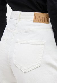 Κοντινό πλάνο ατόμου που φοράει λευκό τζιν με ψηλή μέση, πίσω τσέπες και δερμάτινο ταμπελάκι με την  επιγραφή «VMD» στη μέση.