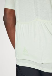 T-shirt de sport vert clair à manches courtes, avec un ourlet froncé et un logo discret. Texture douce et coupe décontractée, associé à un pantalon noir.