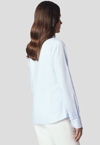 Jasnoniebieska koszula uszyta z gładkiego materiału, z długimi rękawami, zapinanymi mankietami, zaokrąglonym dołem i dopasowanym krojem, pokazana z tyłu.