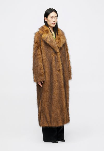 Žena s dlhými čiernymi vlasmi, oblečená v celovečernej hnedej kožušinovej bunde, čiernych nohaviciach a čiernych špicatých topánkach, stojí proti jednému pozadiu.