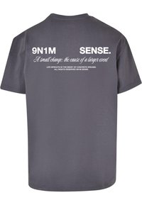 Grå bomull T-skjorte med korte ermer. Bakre del har fet hvit tekst, inkludert "9N1M" og "SENSE," med tilleggsetninger i mindre skrift.