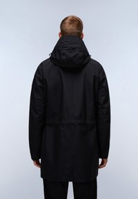 Man med en svart huva lång kappa sedd bakifrån mot en ljus enfärgad bakgrund.