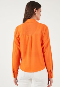Long-sleeve shirt in bright orange, made of lightweight fabric. Features a collar, buttoned cuffs, and a curved hem at the back.
