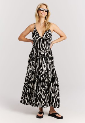 Lachende vrouw die een zwart-wit zebra-prints mouwloze maxi-jurk draagt, zwarte zonnebril, armband en zwarte plateauzandalen, staand met handen in de zij.