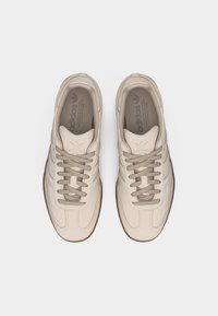 Ζευγάρι μπεζ αθλητικά παπούτσια Adidas με ανοιχτό καφέ κορδόνια και σόλες από καουτσούκ, φωτογραφημένα από πάνω σε λευκό φόντο.