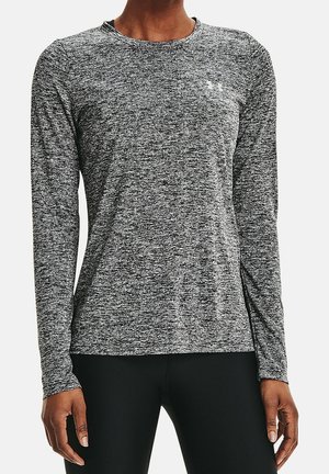 Kobieta nosząca dopasowaną, szarą, melanżową sportową koszulkę z długim rękawem z małym logo na piersi oraz czarne legginsy.