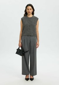 Grijze mouwloze gebreide top met een ronde halslijn, gecombineerd met grijze wijde broek. Model houdt een kleine zwarte handtas vast. Zwarte pumps met spitse neus.