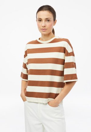 Junge Frau mit braunen Haaren im Pferdeschwanz, die ein lockeres braun-weiß gestreiftes kurzärmeliges Shirt und weiße Hose trägt und die Hände in den Taschen hat.
