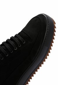Zapatillas de ante negras con suela gruesa de goma negra y marrón con textura estriada, puntera redonda y cordones de cuerda negra pasados por ojales de metal.
