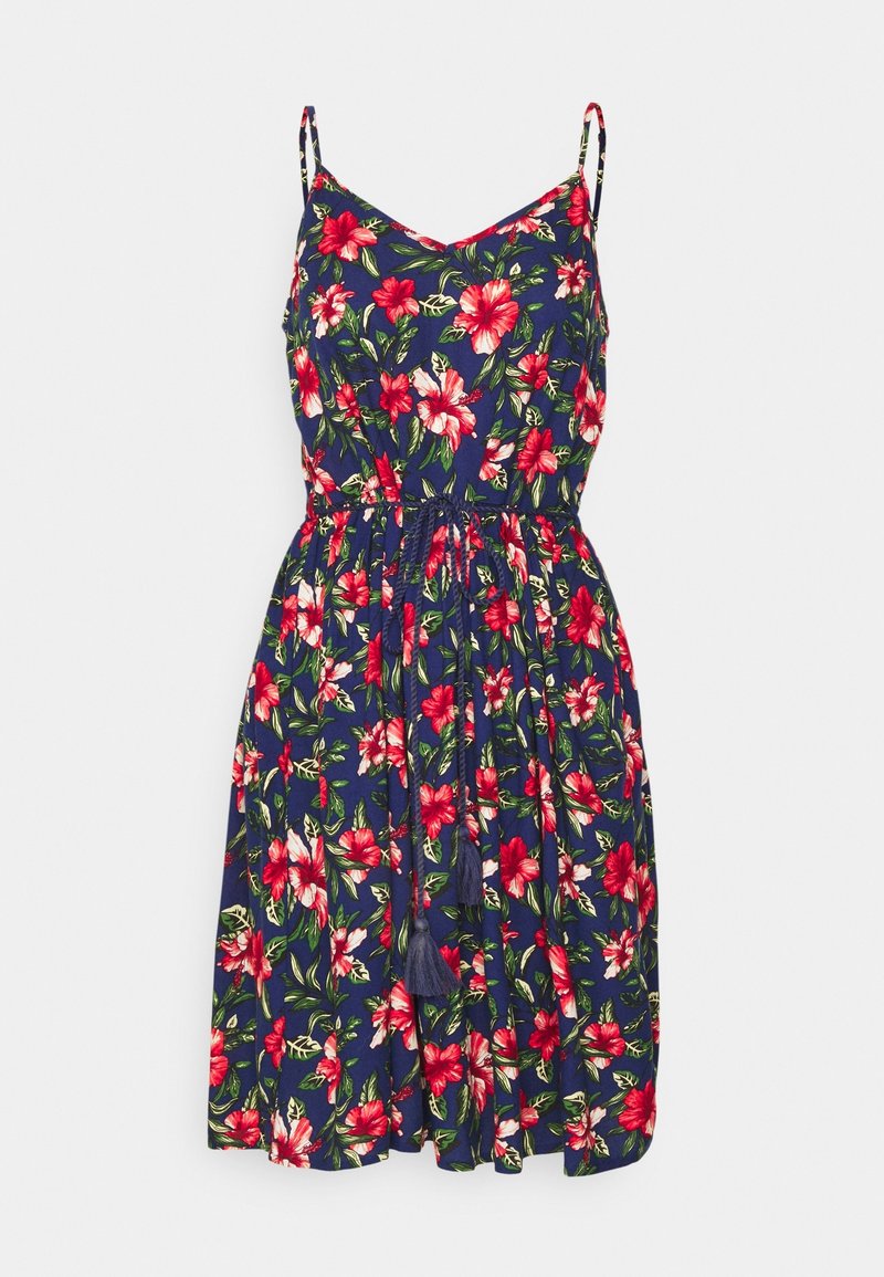 Robe d'été florale en bleu marine avec des hibiscus rouges et des feuilles vertes. Elle possède des bretelles fines, une taille à cordon de serrage et des accents en pompons.