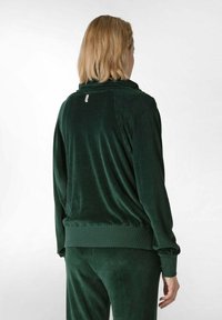 Giacca in velluto verde con colletto alto, polsini e vita a coste. Texture liscia, design semplice e un logo discreto sulla parte posteriore.