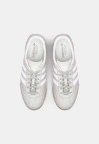 Gaišpelēku zamšadega kedu modelis ar baltām trim svītrām, apaļām auklām un gumijas zoli. Zīmols redzams uz mēles un zoles iekšpusē.