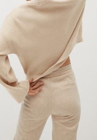Bežový pletený svetr s širokými rukávy, přes který je přehozený odpovídající pár měkkých, přiléhavých kalhot s zadní kapsou a hladkou texturou.