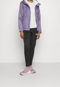 Helllila wasserdichte Jacke mit Kapuze, weißes Hemd darunter, schwarze strukturierte Hose und hellrosa Sportschuhe.