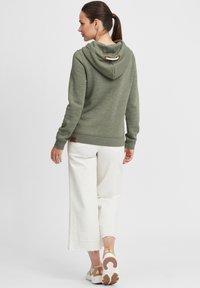 Felpa verde con cappuccio, dalla finitura testurizzata e con un accent loop. Abbinata a pantaloni bianchi a gamba larga e sneaker in tonalità neutre.