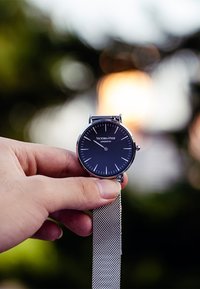 Silberner Mesh-Armbanduhren mit schwarzem Rundzifferblatt, weißen Stundenmarkierungen und Markenlabel. Das Design ist minimalistisch und modern.