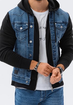 Homme portant un sweat à capuche noir sous un gilet en denim bleu avec des boutons argentés, ajustant un bracelet noir clouté et portant une montre.