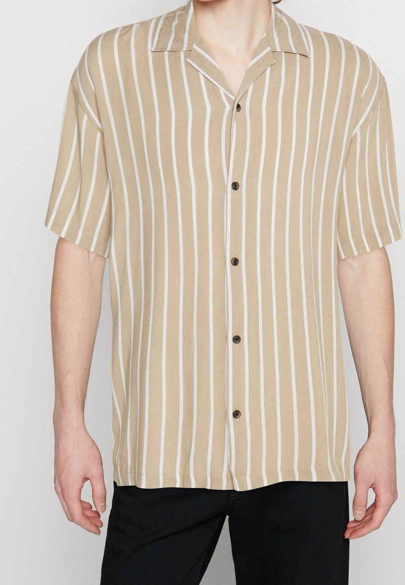 Camicia a maniche corte con bottoni in beige chiaro con righe verticali bianche. Presenta un colletto classico e bottoni neri. Vestibilità casual e trama liscia.