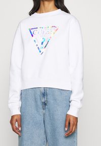 Sweatshirt court blanc avec un logo en triangle holographique, comportant le texte "GUESS U.S.A." et "WASHED". Texture douce et manches longues.
