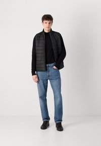 Colete acolchoado preto sobre uma camisola preta de manga longa, jeans de ganga azuis e ténis pretos. Corte reto com costuras e fechos visíveis.