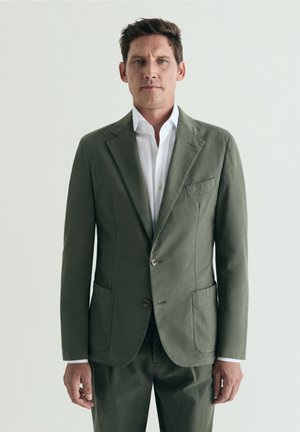 Hombre vestido con chaqueta y pantalones de traje color verde oliva y camisa blanca de vestir, de pie contra un fondo claro y liso, mirando hacia adelante.