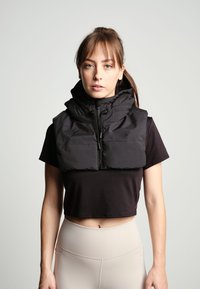 Vestă puffer neagră cu guler înalt, fermoar frontal și șnur. Poartă peste un top scurt negru, asortată cu colanți bej cu talie înaltă.