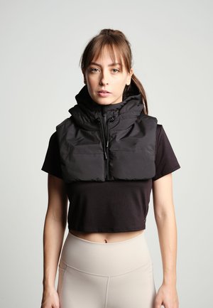Sort puffer vest med høj krave, frontlynlås og snoretræk. Bæres over en sort cropped top, parret med beige højtaljede leggings.