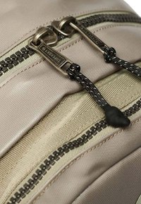 Borsa in tessuto beige con superficie strutturata, dotata di due cerniere argentate, cordini neri e cuciture scure a contrasto lungo le giunture.