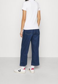 Biała koszulka polo z krótkim rękawem, ciemnoniebieskie szerokie dżinsy, białe sneakersy z czarnymi i czerwonymi akcentami, widok z tyłu.