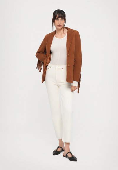 Ruskea mokkajakku, jossa on fringe-yksityiskohtia, valkoiset slim-fit-farkut ja mustat slingback-kengät. Yksinkertainen toppi alla, yleisilmeeltään neutraali väripaletti.