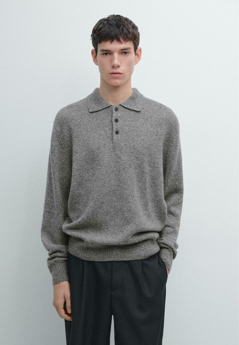 Szary dziergany sweter z kołnierzem i plisą na trzy guziki. Posiada długie rękawy i luźny krój. Całość dopełniają ciemne spodnie.