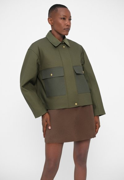 Olive green cropped jacket with two front flap pockets, gold buttons, a collar, and soft fabric texture, paired with a brown mini skirt.
