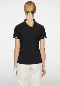 Svart polo-tröja med korta ärmar, vita accentstrimmor på ärmarna och en klassisk krage. Har en mjuk, stretchig tygkonstruktion för komfort.