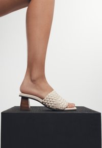 Sandalia de pantalla tejida en color crema, con punta cuadrada y tacón de madera cilíndrico, exhibida sobre una plataforma negra contra un fondo claro.